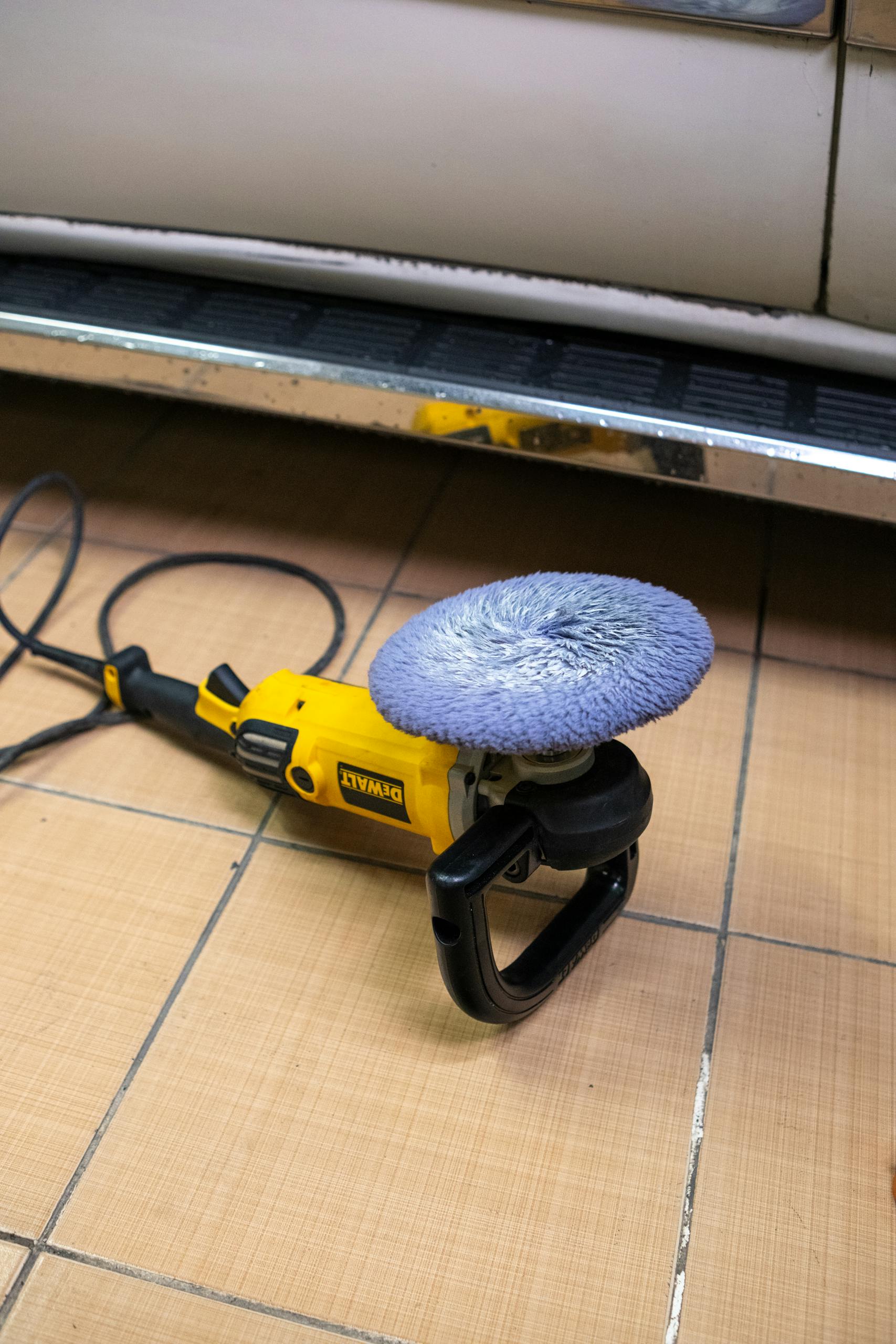 A yellow electric car polisher resting on a tiled garage floor, next to a vehicle.